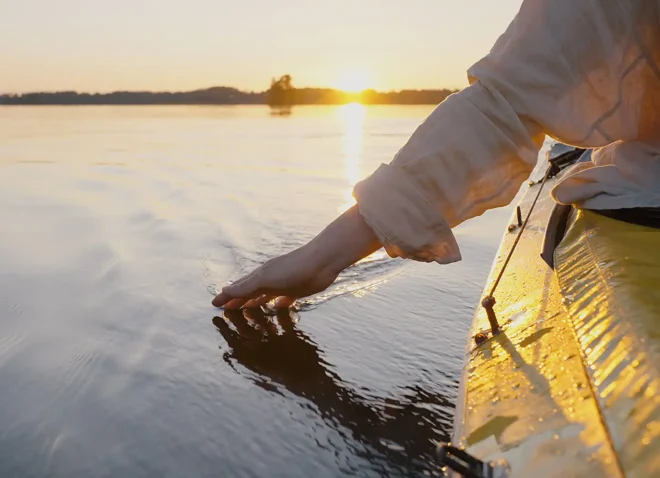 Klimafreundlich Reisen, ein gelbes Kanu auf einem See beim Sonnenuntergang am Horizont, eine Hand der Kanufahrerin berührt die Wasseroberfläche.