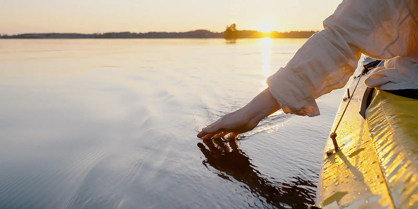 Climate-friendly travel, a yellow canoe on a lake at sunset on the horizon, a canoeist's hand touching the surface of the water.